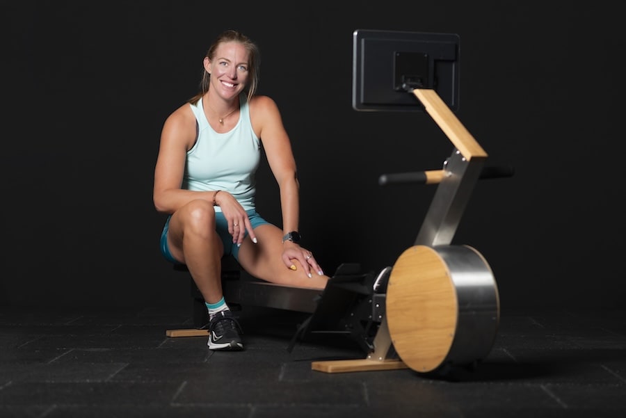 Femme souriante assise sur un rameur avec un écran devant elle, portant un débardeur bleu et un short.