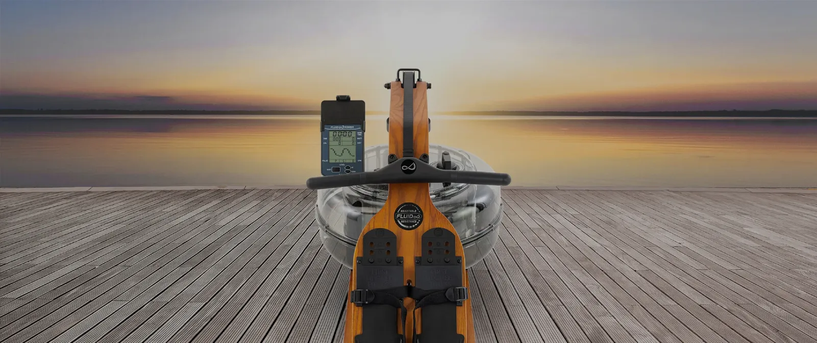 Rameur Fluid Rower Viking en bois orange avec écran numérique, posé sur une plateforme au bord d’un lac au coucher du soleil, appareil d’entraînement