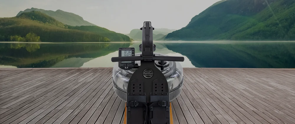 Rameur Apollo Plus Bluetooth noir et gris avec écran numérique, posé sur une terrasse en bois au bord d’un lac entouré de montagnes.