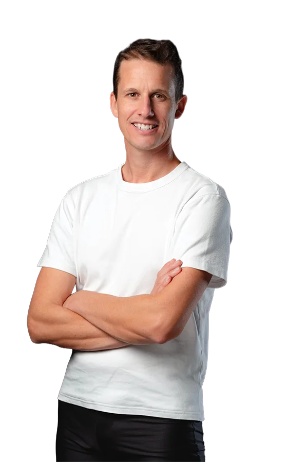 Portrait de William Coach avec t-shirt blanc et pantalon sombre, bras croisés, sourire, cheveux courts bruns, fond sombre contrasté.