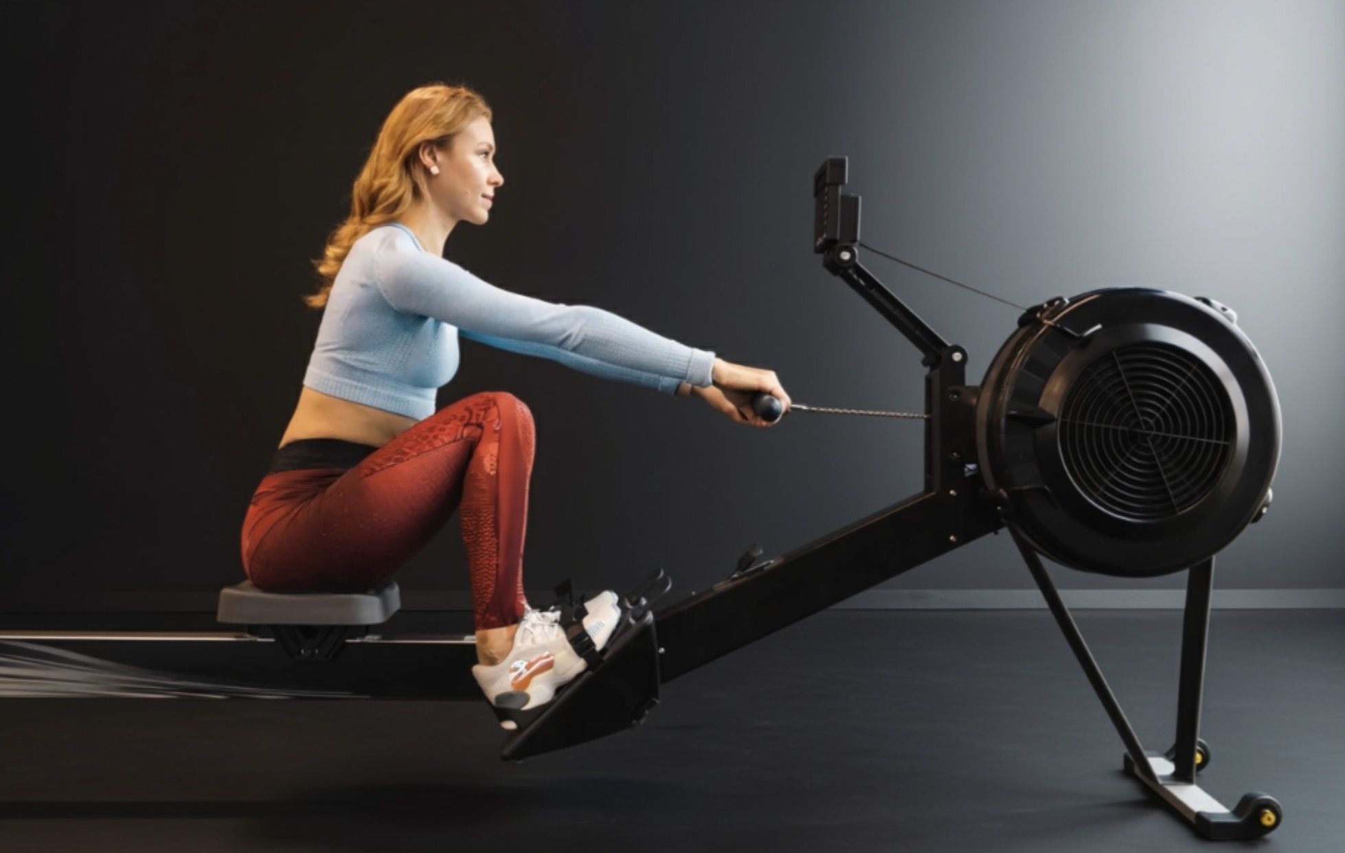 Assise sur un rameur, une sportive aux cheveux roux tient la poignée, genoux fléchis et pieds calés sur les supports.