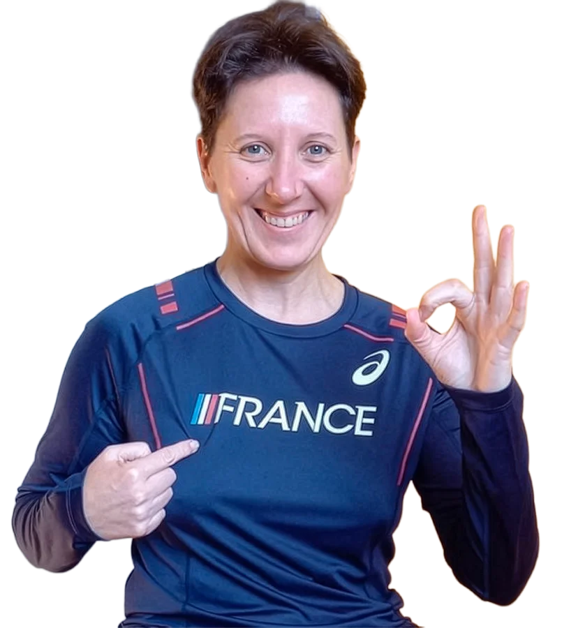 Souriante, Bénédicte porte le maillot bleu de l’équipe de France, en désigne l’inscription et forme un geste OK de l’autre main.
