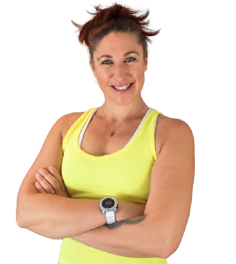 Une femme souriante aux cheveux roux attachés porte un débardeur jaune et une montre de sport, les bras croisés.